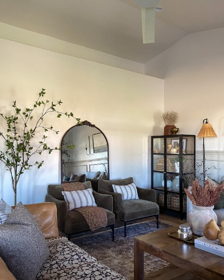 A cozy living room featuring two comfortable armchairs with striped cushions, a wooden coffee table, and a decorative floral arrangement. A tall plant stands beside a full-length mirror, and a stylish storage cabinet is also present, adding warmth and charm to the space.