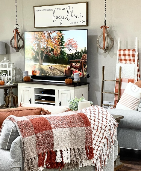 Cozy living room featuring a warm, autumn-inspired atmosphere with a red and beige checkered throw blanket, a display of seasonal decor, and a scenic fall image on the TV.