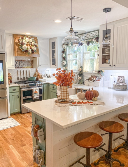 A cozy kitchen adorned with autumn decor, featuring a vase of fall foliage, decorative pumpkins, and warm lighting. The space combines rustic elements with modern conveniences, creating an inviting atmosphere.