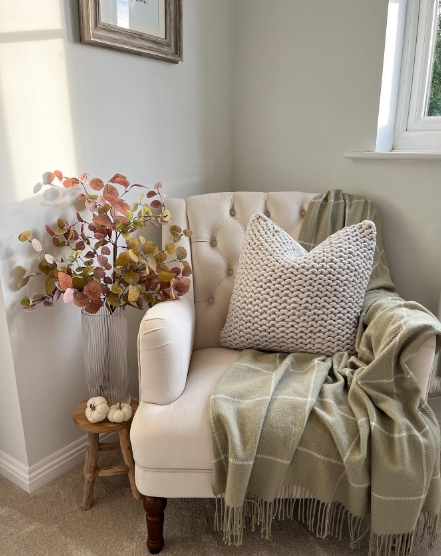 A cozy corner featuring a light-colored armchair adorned with a textured knit pillow and a soft green throw. Next to it, a small wooden stool holds decorative pumpkins, while a vase with autumn-hued foliage adds a seasonal touch.