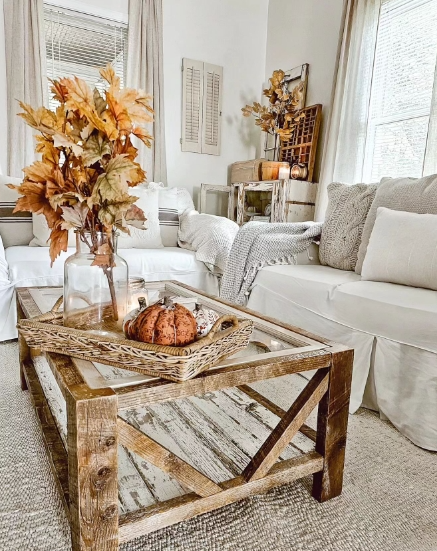 A cozy living room featuring a rustic wooden coffee table with a glass top. The table is adorned with a decorative basket holding small pumpkins, and a vase filled with autumn leaves. Plush white sofas with soft pillows create a warm and inviting atmosphere.