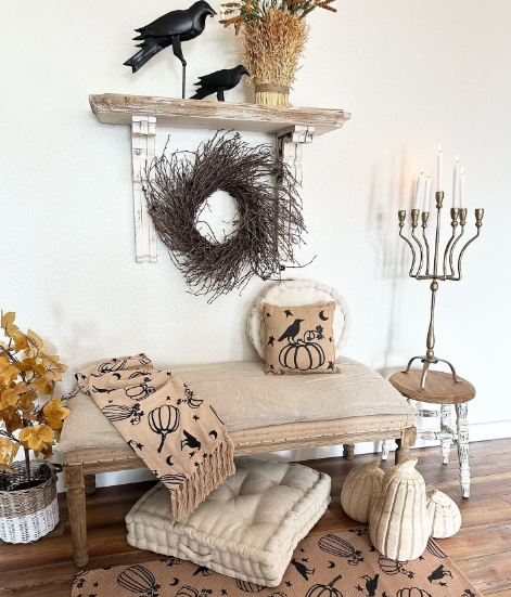 A cozy entryway featuring a bench with a pumpkin-themed pillow, soft mats, and a decorative wreath on the wall. Accents include candles, seasonal decor, and decorative pumpkins alongside dried foliage.