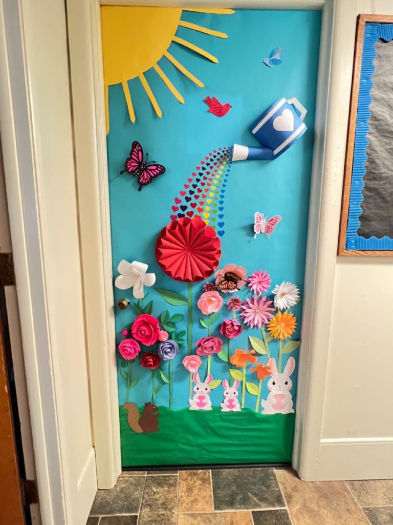 Bright and cheerful spring classroom door with a giant watering can pouring love onto blooming flowers.