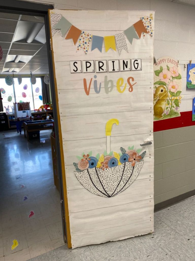 Minimalist farmhouse-style spring classroom door with pastel banners and an umbrella full of flowers.