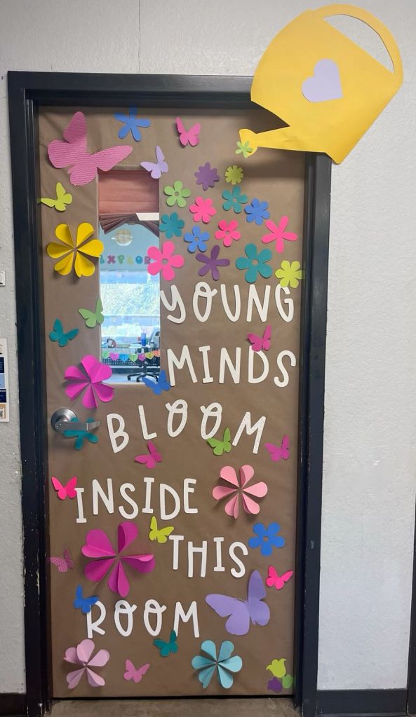 Garden-themed classroom door featuring a watering can pouring colorful flowers and butterflies.