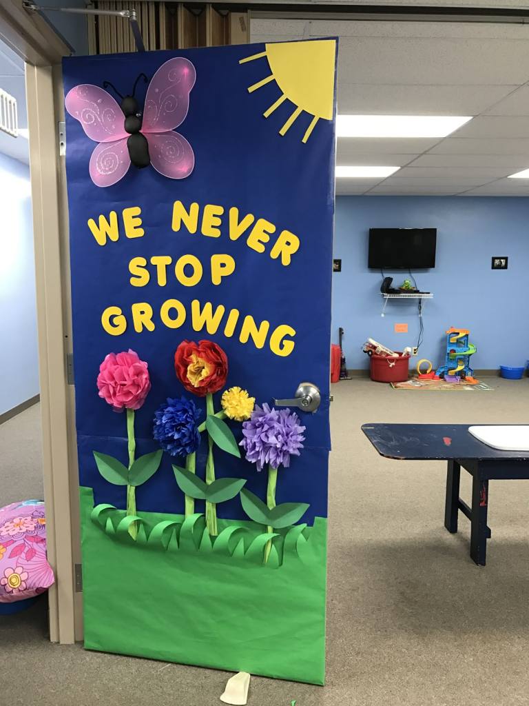 3D paper flower classroom door with the phrase 'We Never Stop Growing' and a large pink butterfly.