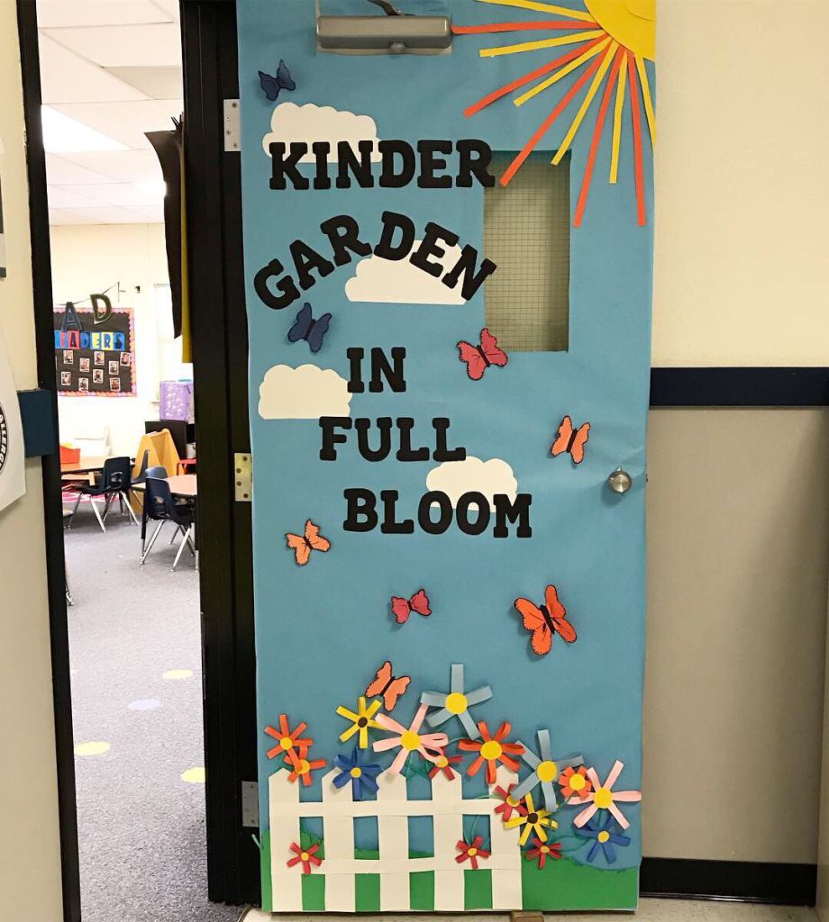 Kindergarten classroom door decorated with butterflies, a white picket fence, and 3D spring flowers.