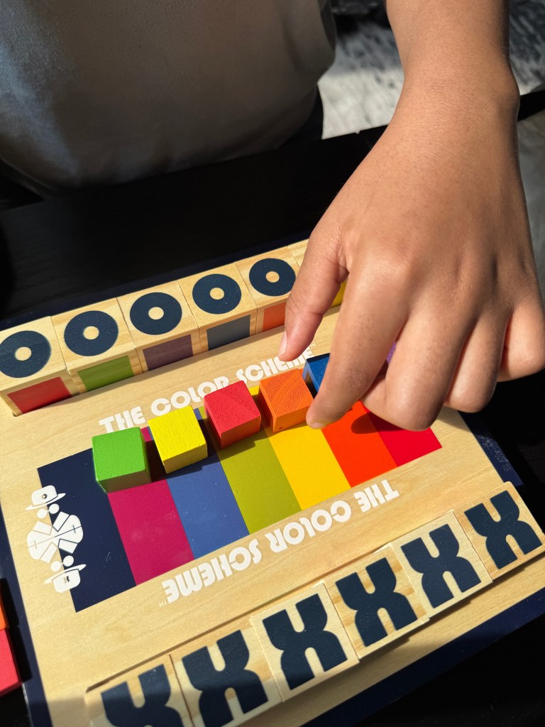 A child's hand reaching for a colored block on 'The Color Scheme' board game, featuring vibrant colors and wooden pieces arranged on a game board.