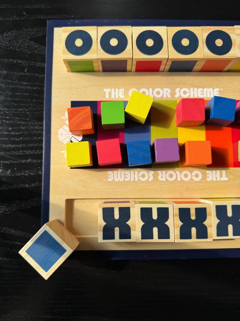 A colorful game board for 'The Color Scheme' featuring vibrant wooden blocks in bright colors and code cubes marked with 'X' and 'O'.