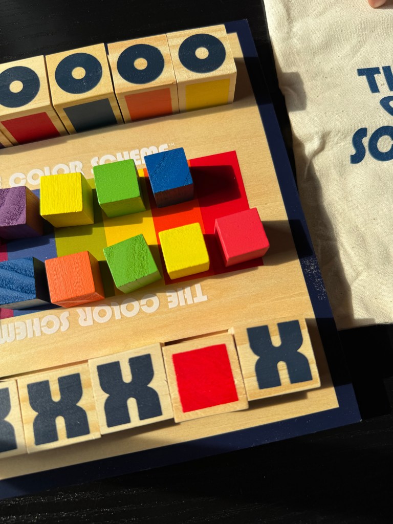 A close-up view of 'The Color Scheme' board game featuring colorful wooden blocks in various shapes, a wooden game board with the game's title, and a soft canvas storage pouch nearby.