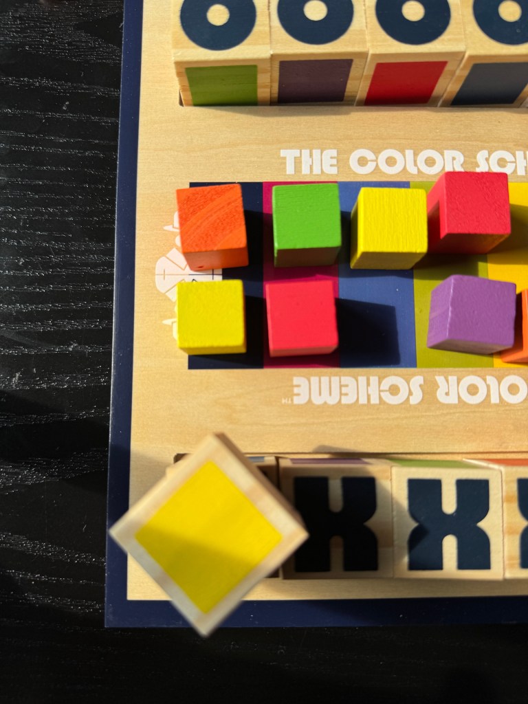 Close-up view of the Color Scheme board game, featuring colorful wooden blocks in various shades, including red, green, yellow, and pink, arranged on a game board with 'THE COLOR SCHEME' printed in white.