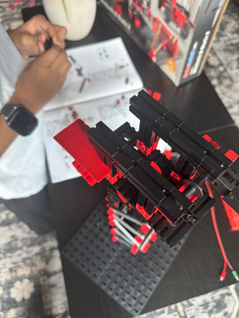 Close-up view of a child assembling a red and black Fischer Technik STEM kit model, with building instructions visible in the background.
