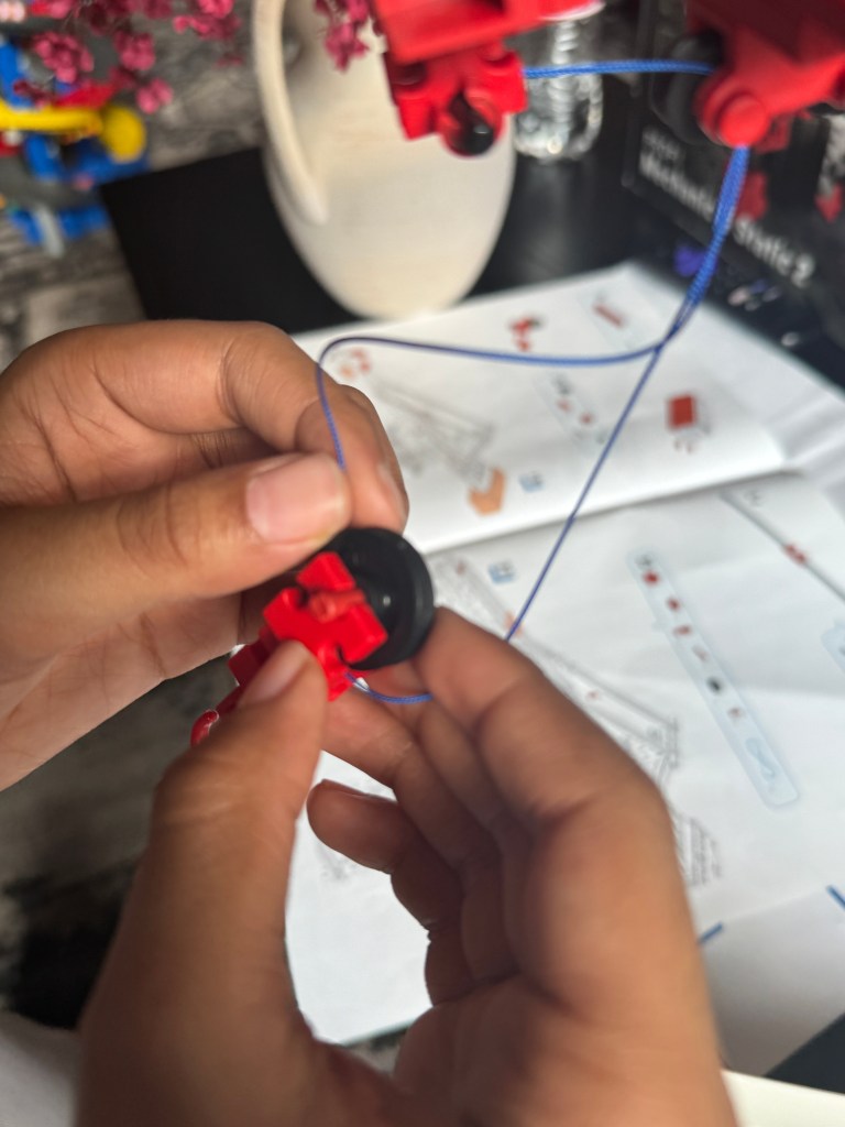 A close-up of a child's hands assembling a component of the Fischer Technik STEM Kit, featuring a red piece connected to a black wheel with a blue string, with an instruction manual in the background.