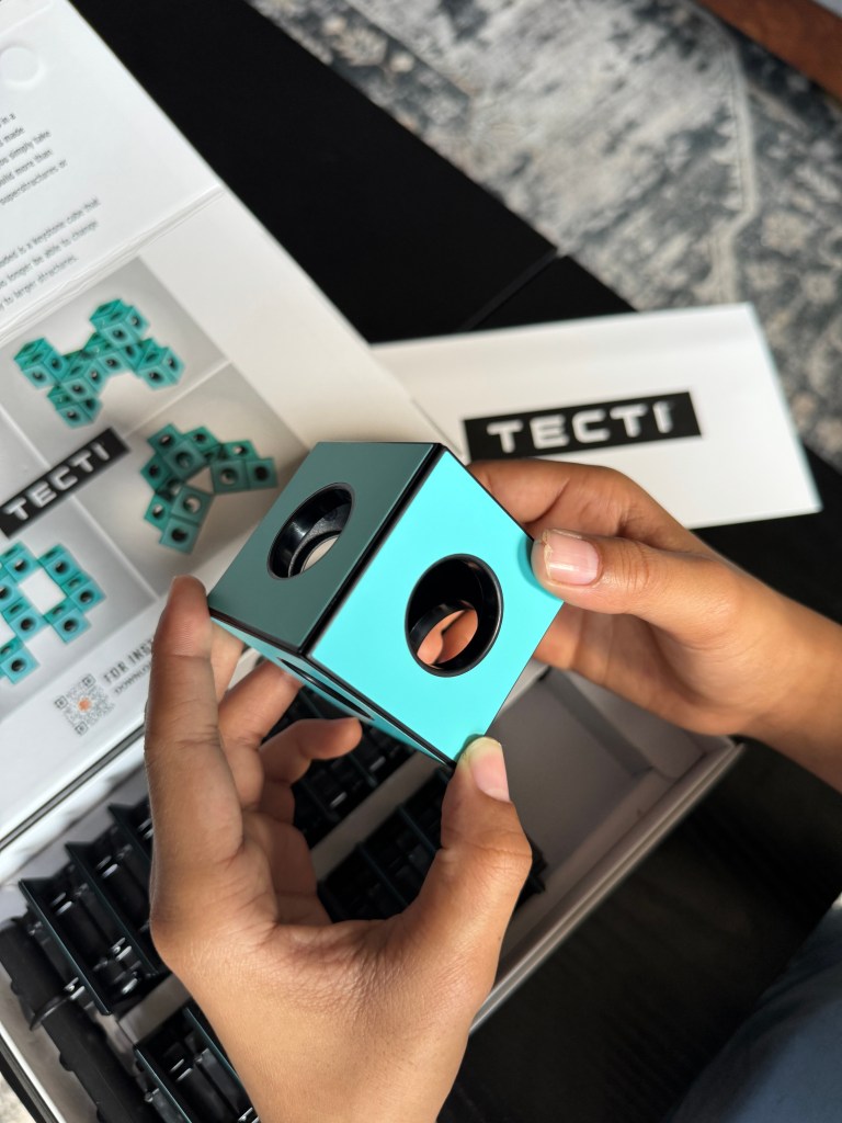 A person holding a turquoise building cube from the Tecti kinetic building system, with a neatly organized box containing additional pieces visible in the background.