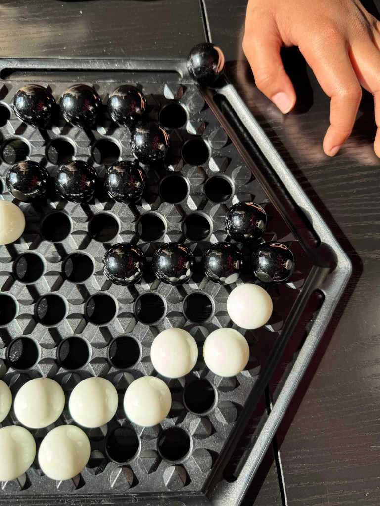 Close-up of an Abalone game board featuring black and white marbles, with a hand positioned near a black marble ready to slide.