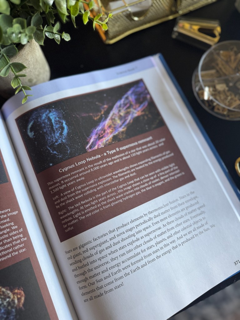An open book showing the Cygnus Loop Nebula illustration with descriptive text about supernova remnants, alongside a potted plant and various desk items.
