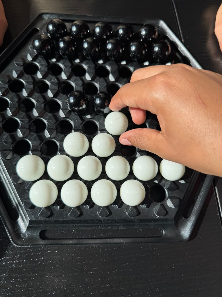 A hand reaching to move a white marble on a hexagonal board featuring black and white marbles, set against a dark wooden surface.