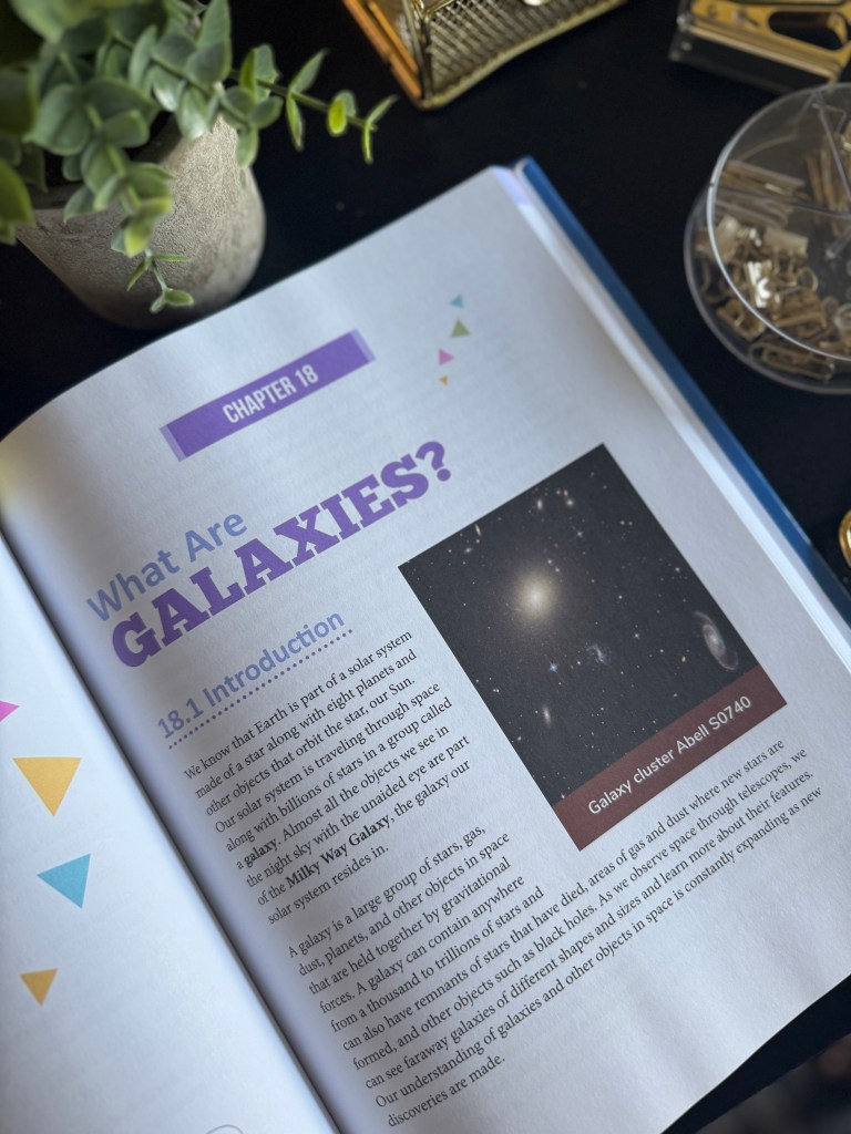 A close-up of an open science textbook page discussing galaxies, with a vibrant photo of a galaxy cluster and decorative elements like colorful shapes on the page.