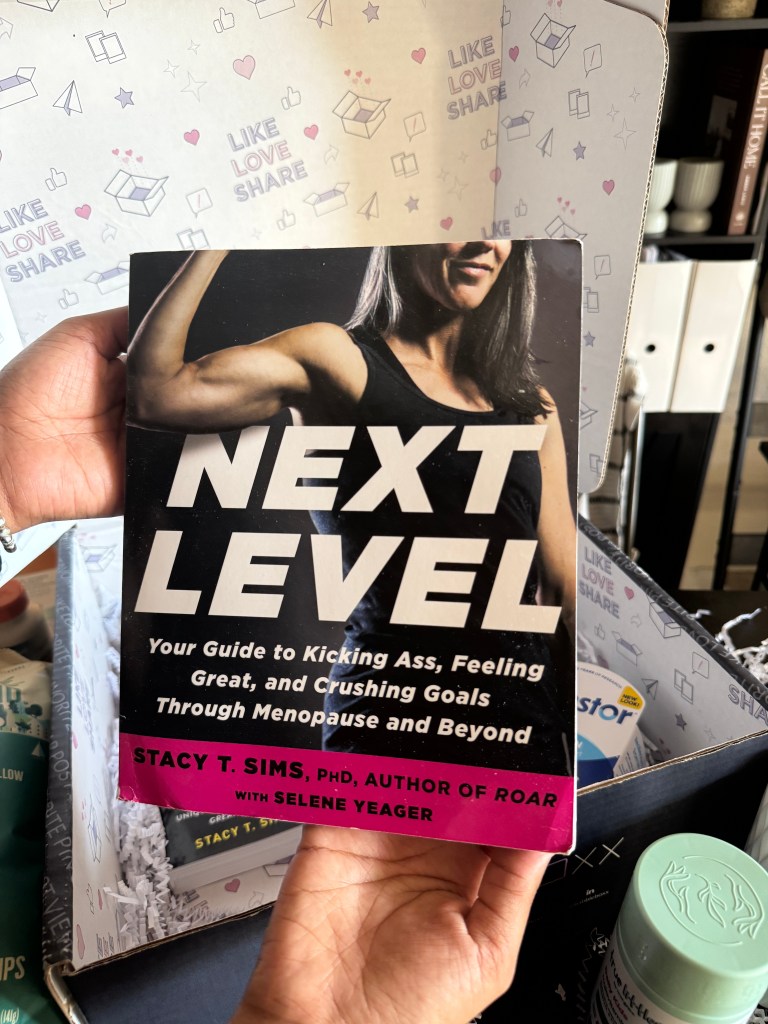 A hand holding the book 'Next Level' by Stacy T. Sims, with a background featuring a patterned box and wellness products.