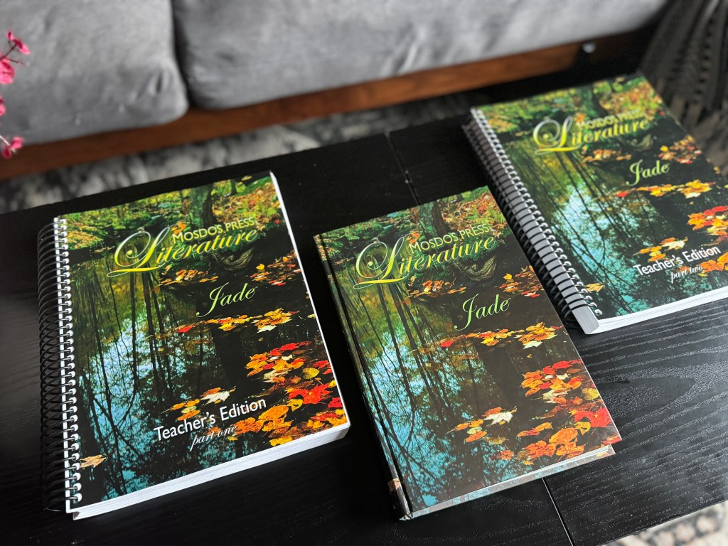 Three books spread out on a dark wooden table, featuring the titles 'Mosdos Press Literature: Jade' and 'Teacher's Edition' with a serene forest image on the cover.