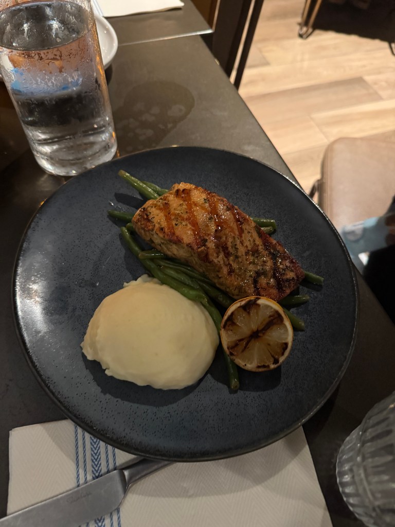 A plated dish featuring grilled salmon fillet, sautéed green beans, and creamy mashed potatoes, garnished with a lemon slice, served on a dark plate next to a glass of water. Fireside Restaurant at Great Wolf Lodge in Grapevine, TX. 
