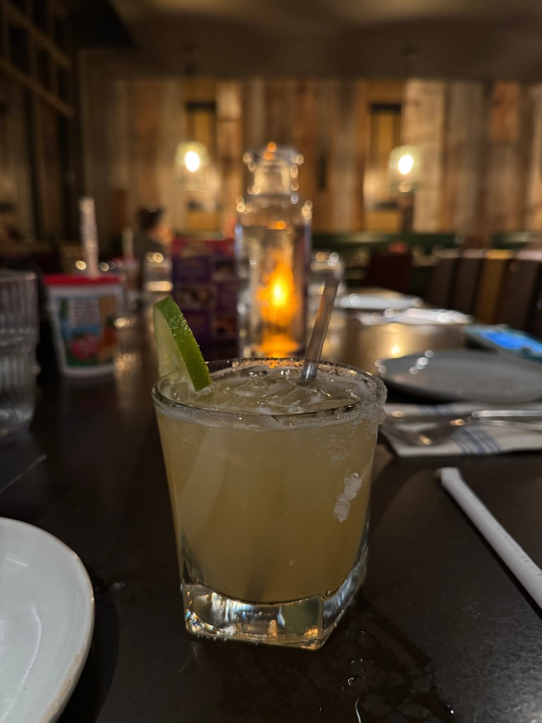 A close-up image of a margarita glass filled with a yellowish drink, garnished with a lime wedge. The glass features a salt rim and is placed on a dark table, with blurred background elements including a water pitcher and other dining items. Fireside Restaurant at Great Wolf Lodge in Grapevine, TX. 