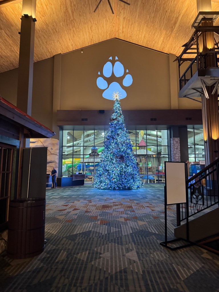 Indoor lobby of Great Wolf Lodge featuring a large, decorated Christmas tree in blue lights, a paw print projection on the wall, and view of the water park through glass doors.