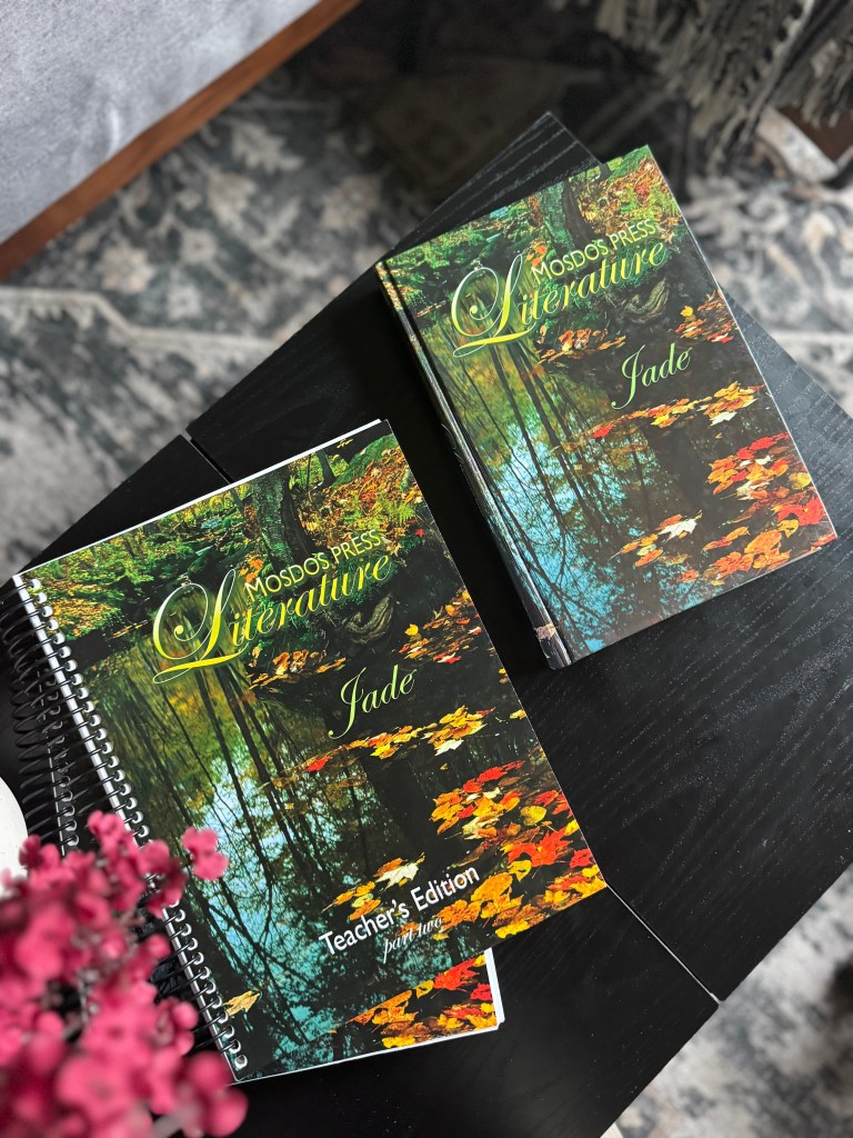 Two Mosdos Press Literature: Jade books on a wooden table, one being a Teacher's Edition, surrounded by flower decor.