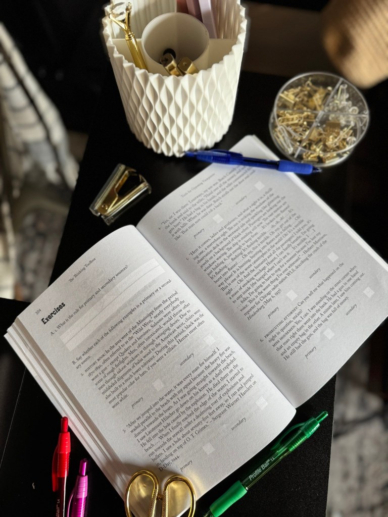An open book titled 'The Thinking Toolbox' with exercises visible, placed on a dark surface surrounded by various writing utensils, including colorful pens and paper clips, alongside a decorative pen holder.