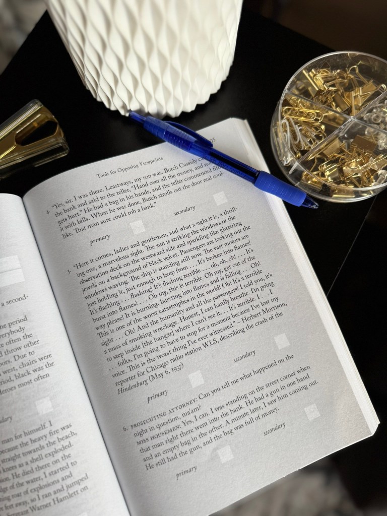 Close-up of an open book page with text focused on tools for opposing viewpoints, a blue pen, a gold paperclip holder, and a textured white lamp in the background.