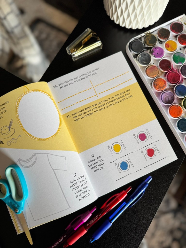 An open art book featuring prompts and illustrations, placed on a table alongside scissors, colored pens, and a watercolor palette.
