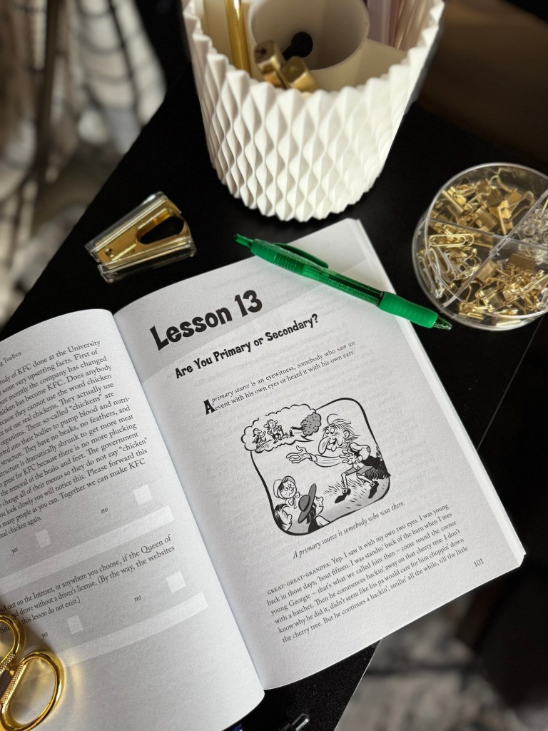 An open book displaying Lesson 13 titled 'Are You Primary or Secondary?' with text and illustrations, accompanied by a green pen, a stapler, and a container of office supplies on a dark surface.