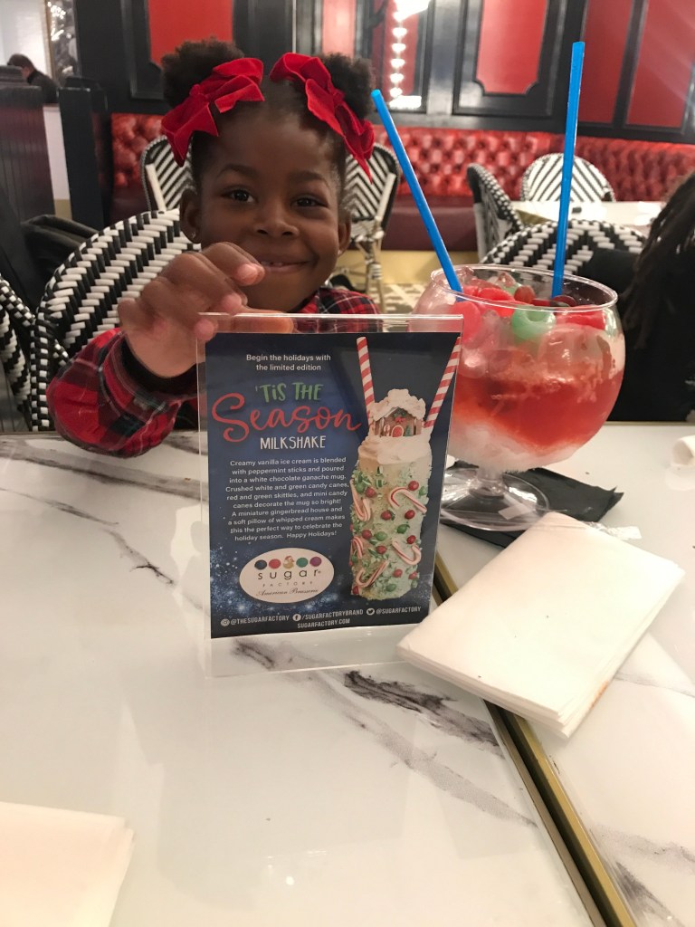 Young girl holding Sugar Factory Biloxi's seasonal holiday menu sign featuring the limited-edition Christmas milkshake.