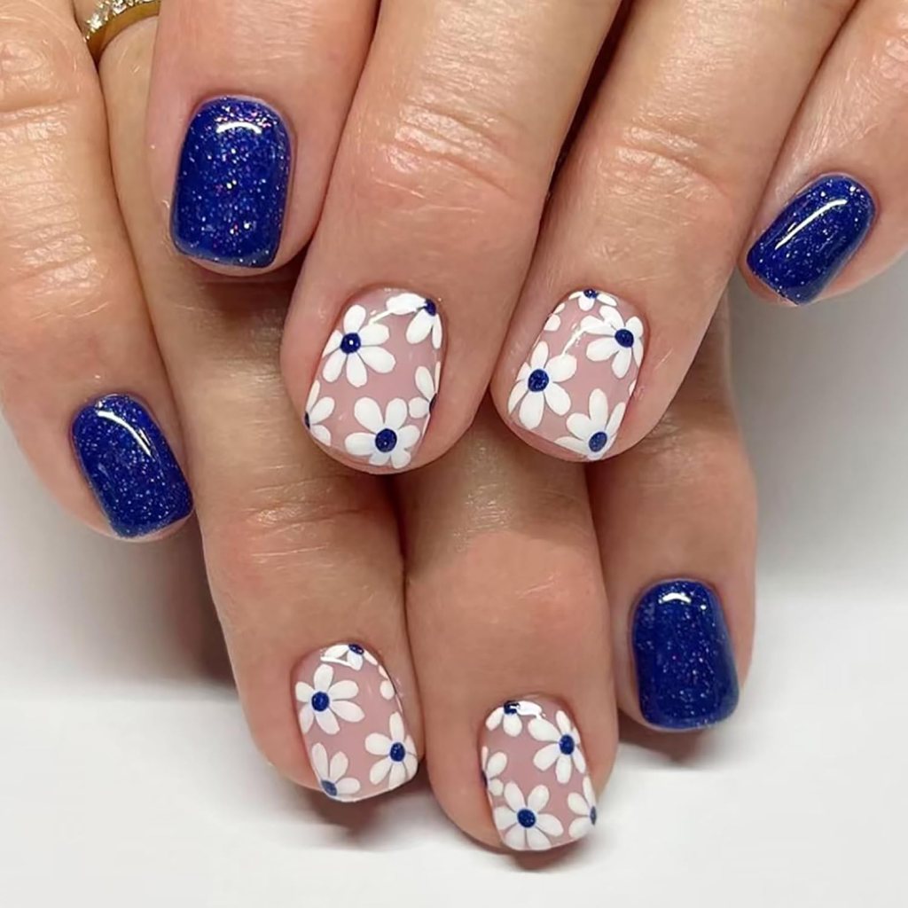 Close-up of manicured hands with alternating nail designs: two fingers feature a deep blue glitter polish, while the other fingers have a light pink base with white daisies and blue centers.