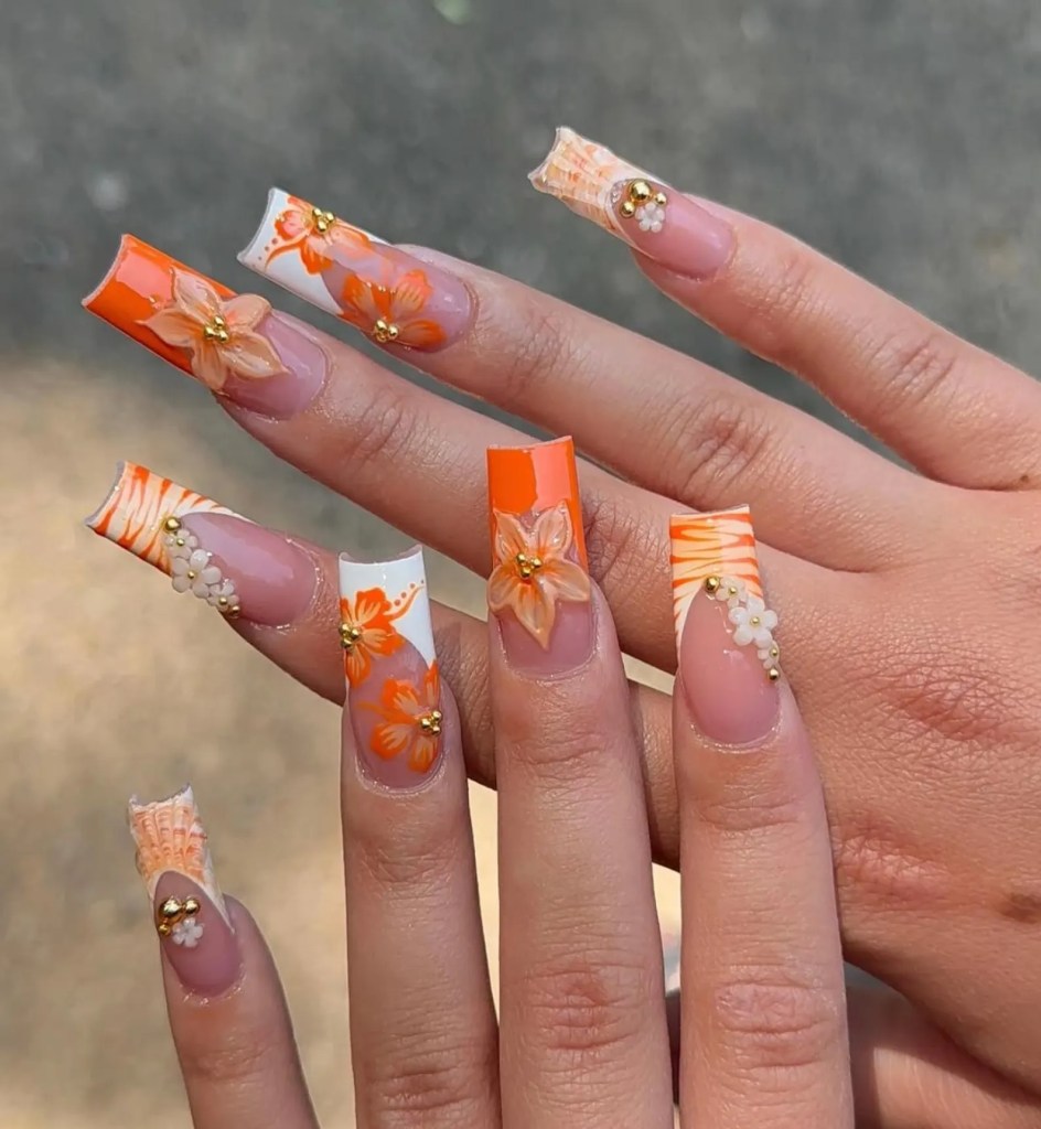 Long acrylic nails with orange polish, floral nail art, and rhinestone details
