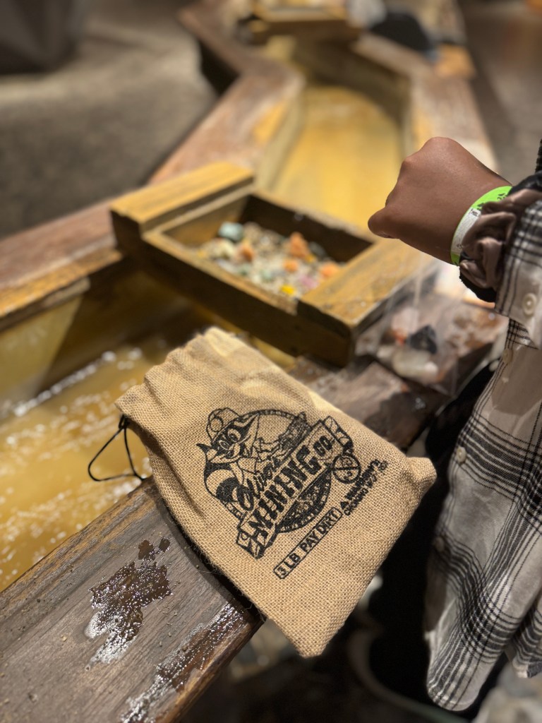 A child mining for gemstones at Oliver's Mining Co. in Great Wolf Lodge, using a sifting tray and holding a burlap bag with the logo.