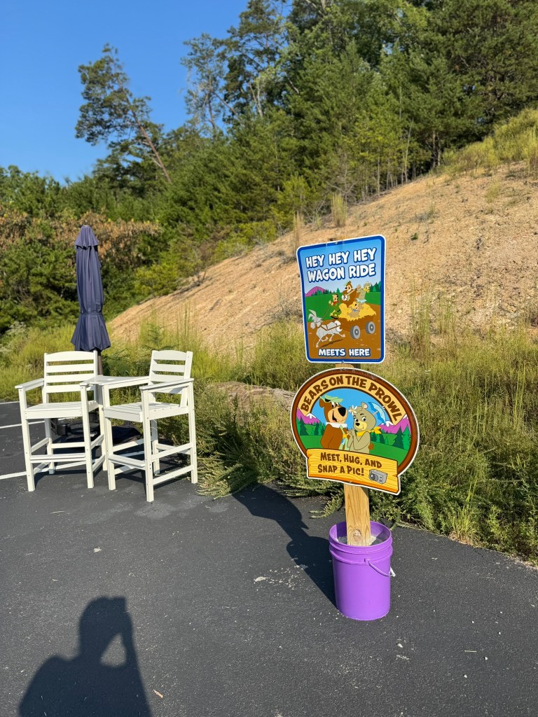 A sign for a wagon ride reading 'Hey Hey Hey Wagon Ride Meets Here' and a colorful sign for 'Bears on the Prowl' with instructions to 'Meet, Hug, and Snap a Pic!' On the side, there are two white chairs and a purple container.