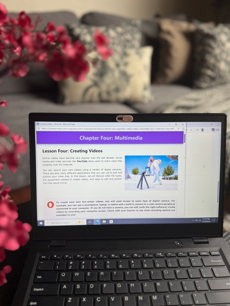 A laptop displaying a lesson on creating videos from a tech curriculum, with a floral arrangement in the foreground and a cozy living room background.