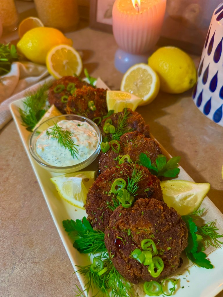 A platter of crispy salmon patties garnished with green onions and fresh herbs, accompanied by a bowl of creamy dip and lemon wedges.