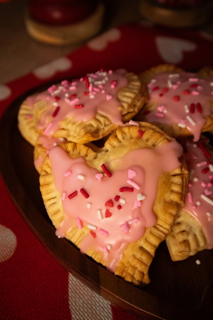 A plate of heart-shaped pastries topped with pink icing and colorful sprinkles, placed on a red tablecloth with white hearts.