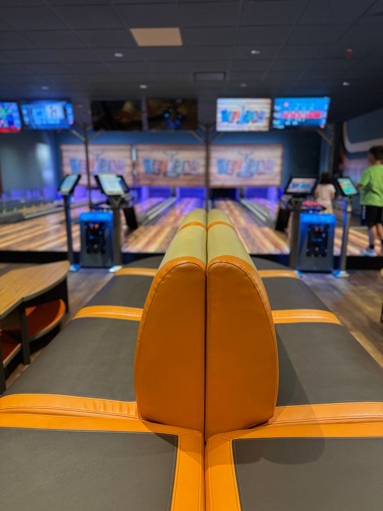 Interior view of Ten Paw Bowling at Great Wolf Lodge, featuring bowling lanes and video screens, with a focus on a seating area in the foreground.