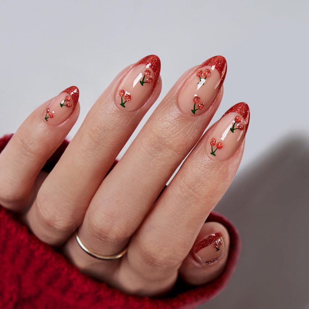 A close-up of a hand with manicured nails featuring a nude base, red glitter tips, and hand-painted floral designs with small red flowers and green stems.