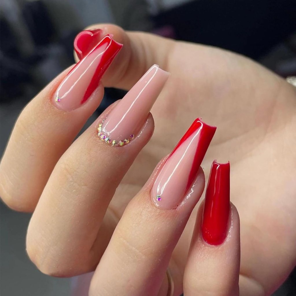 Close-up of a hand showing stylish long nails with a combination of red and nude polish, featuring a decorative rhinestone detail.