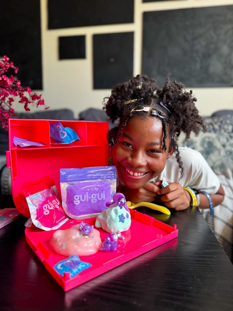 A cheerful child with curly hair smiles while leaning over a pink playset filled with colorful slime and small figurines. The background is a cozy living space.