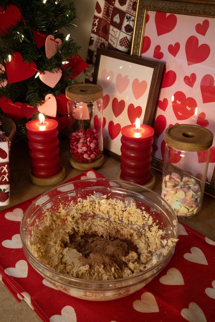 A bowl of cookie dough with cocoa powder on top, surrounded by festive decorations including a small Christmas tree adorned with heart shapes, candles, and jars filled with colorful candies, on a heart-patterned tablecloth.