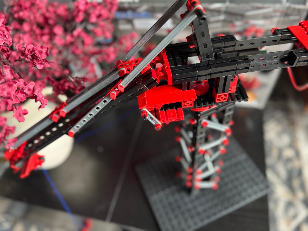 A close-up view of a red and black Fischer Technik crane model, showcasing its intricate construction and engineering features among decorative pink flowers.