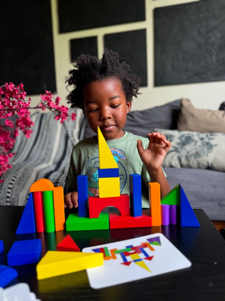 A child playing with colorful geometric building blocks on a table, constructing a structure. There are also vibrant flowers in the foreground and a card with a pattern nearby.