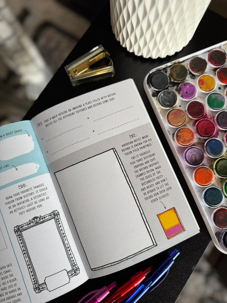 An open art book with prompts and blank spaces for drawing, accompanied by watercolor paints and pens, placed on a dark surface.