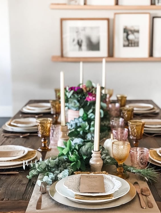 A beautifully styled Thanksgiving table setting featuring layered dinnerware, elegant glassware, and floral elements. The table is adorned with candle holders and greenery, creating a warm and inviting atmosphere.
