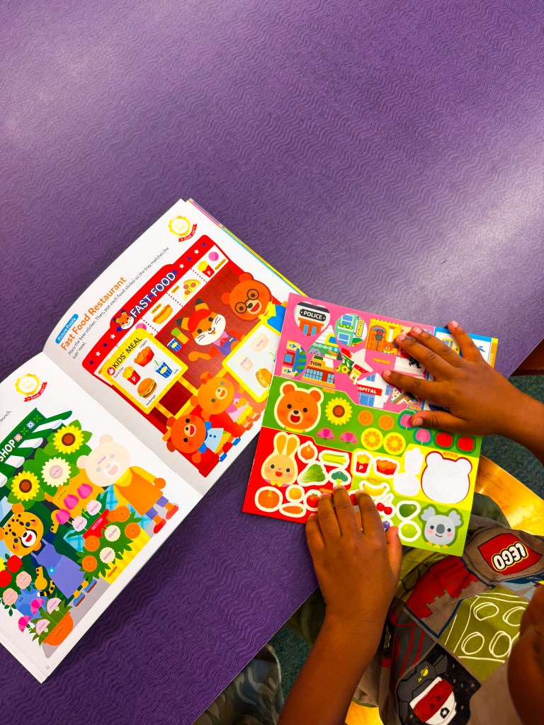 A child’s hands holding colorful sticker sheets and a themed activity book on a purple table, featuring cartoon characters and scenes.